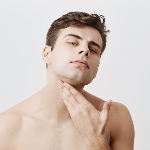 Confident naked handsome man with pure and healthy skin, stylish haircut, blue eyes, keeps hand on chin, checking how good he is shaved, isolated against gray background. People and lifestyle concept.